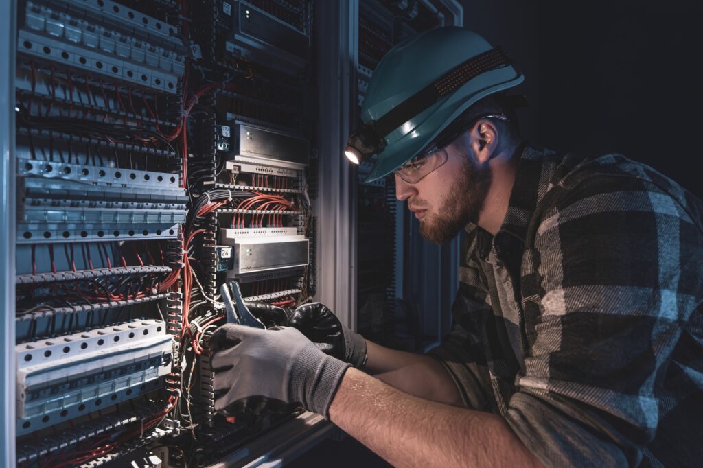A male electrician works in a switchboard in overalls against the backdrop of emergency lighting.